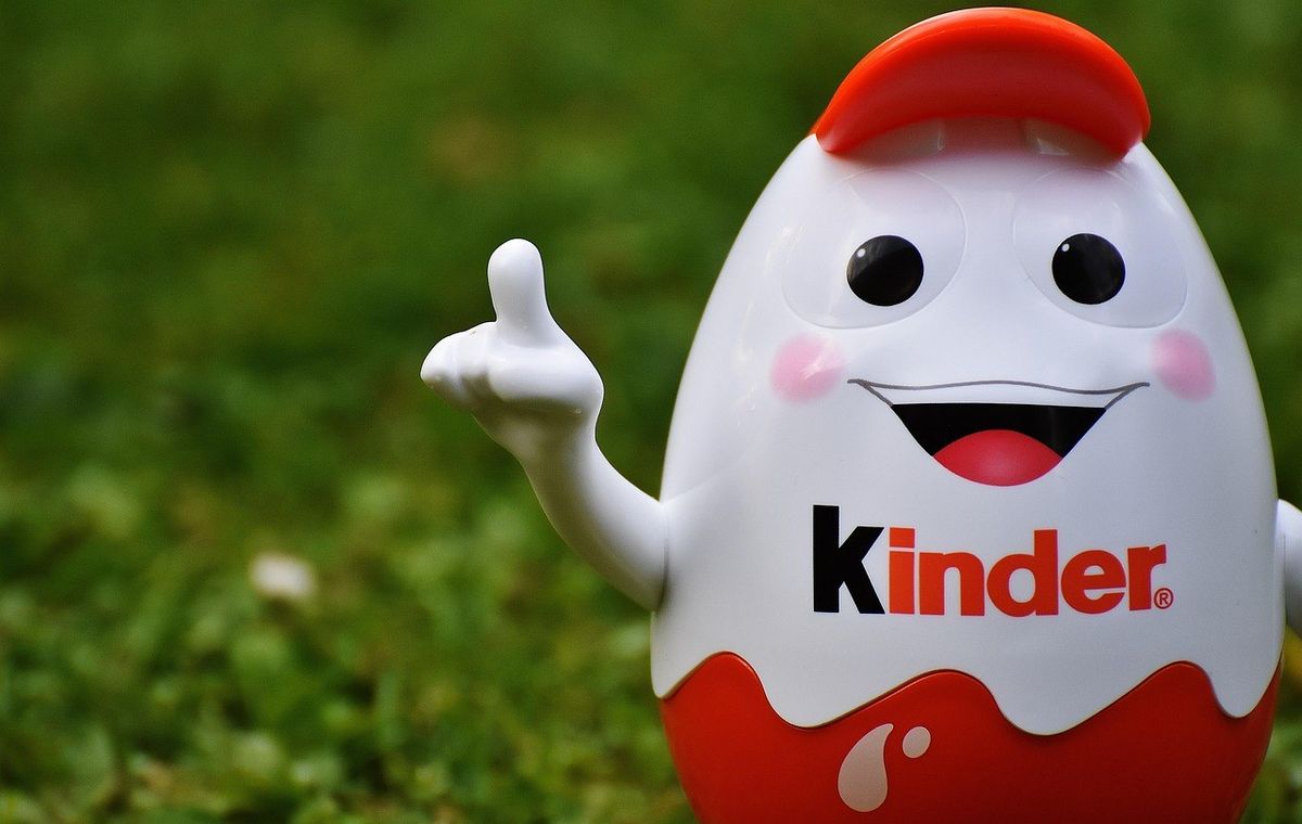Oiseau Kinder souriant, chapeau rouge, signe de paix, logo visible, herbe en arrière-plan